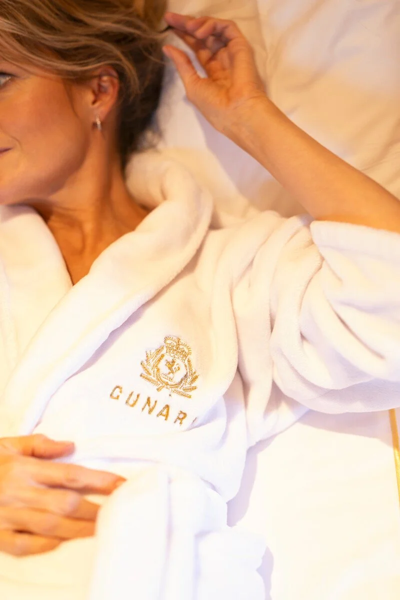 A woman lying on a bed wearing a white bathrobe with an embroidered crest and the word "GUNAR" on it, smiling and resting with her hand near her head.