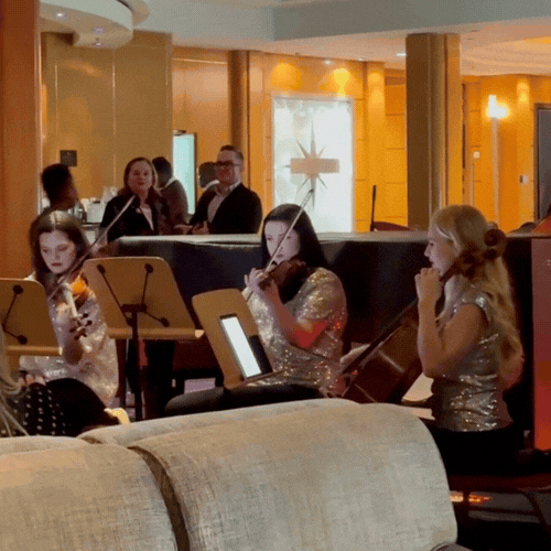 Three young girls in sequined dresses playing violins in a living room or lounge area, with a group of adults in the background, some of whom are seated and engaging in conversation.