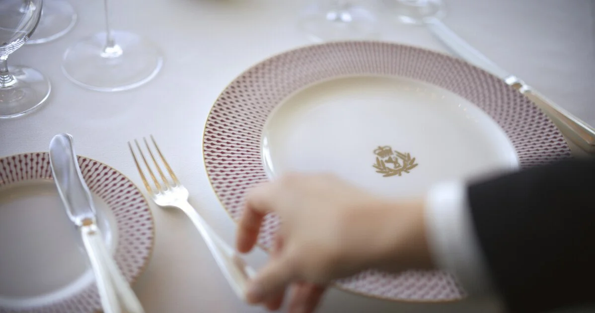 Elegant dinner table setting with white and red patterned plates, silver forks, knives, and wine glasses on a white tablecloth.
