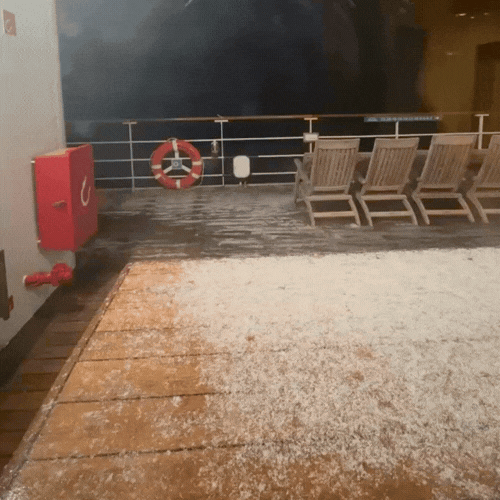 Deck of a cruise ship with wooden lounge chairs, safety equipment including a life ring and fire extinguisher, and an ocean view at night.