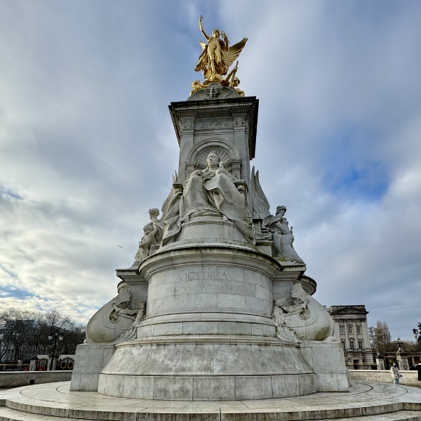 A tall monument with intricate carvings and statues, topped with a gold statue of a winged figure, set against a cloudy sky.