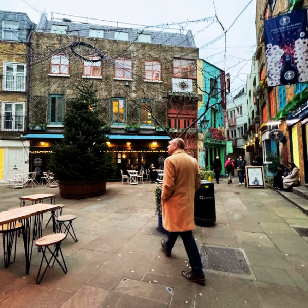 A man in a tan coat walking in a colorful outdoor city square with café seating, a large Christmas tree, and buildings decorated with string lights and street art.