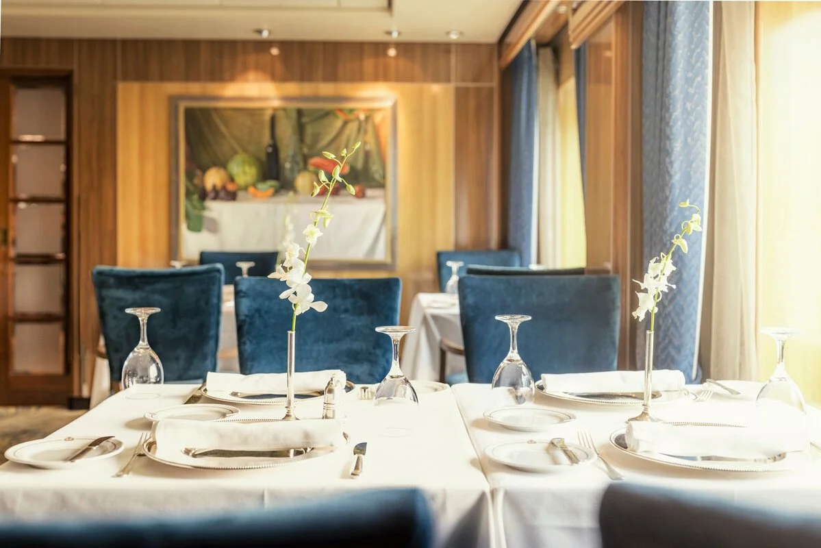 Elegant dining table set with white tablecloth, plates, glasses, napkins, and flowers in the foreground, with blue chairs and a framed painting on the wooden-paneled wall in the background, illuminated by natural light from large windows with curtains.