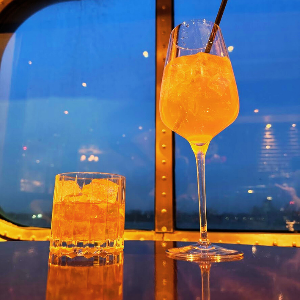 A glass of orange cocktail with ice and a straw and a short glass with ice and an orange drink on a table, with a dark blue sky and city lights in the background.