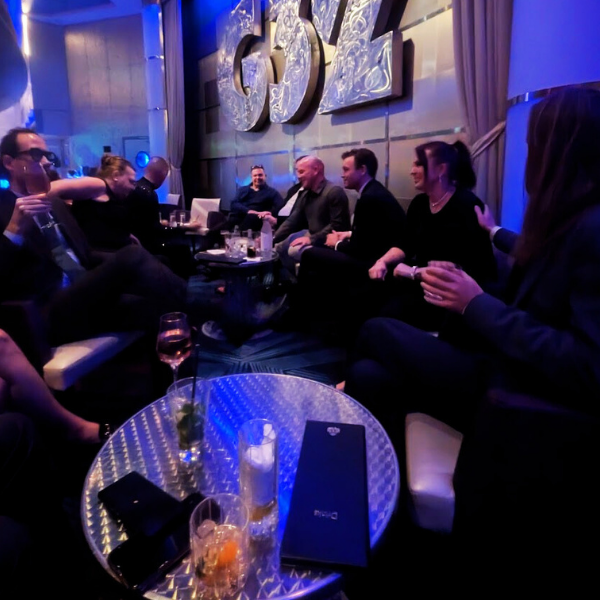 Group of people sitting and chatting in a dimly lit lounge with large 3D letters on the wall behind them and drinks on the table in front.