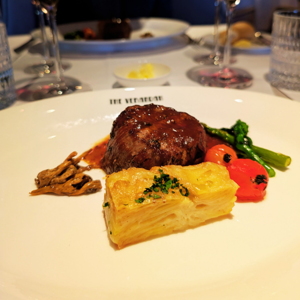 A plated gourmet meal featuring a beef or meatloaf with gravy, a slice of layered potato gratin, sautéed mushrooms, and green beans with cherry tomatoes garnished with herbs, on a white plate at a restaurant.