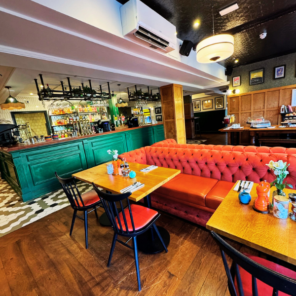 Interior of a cozy restaurant with a red tufted bench, wooden tables, black chairs with red cushions, a teal bar counter, and a well-stocked bar in the background. Decor includes framed pictures on the wall and small decorative items on the tables.