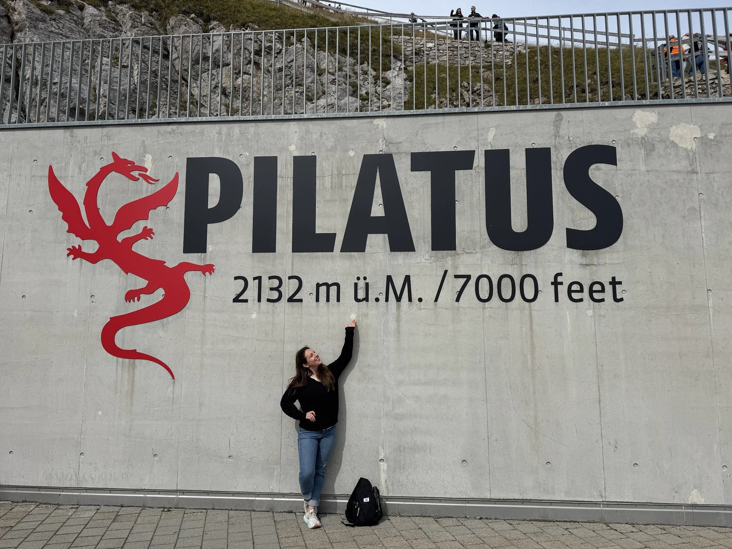A woman standing with one hand on her hip and the other pointing up, smiling in front of a large sign that reads 'PILATUS' with an illustration of a red dragon. The sign mentions an elevation of 2132 meters or 7000 feet. The background shows a hillside with rocky terrain and a metal railing, with some people sitting and standing above.