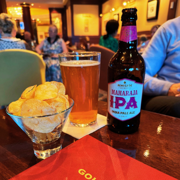 A glass of beer, a bowl of potato chips, and a bottle of Maharaja IPA on a wooden table in a restaurant or pub with people in the background.