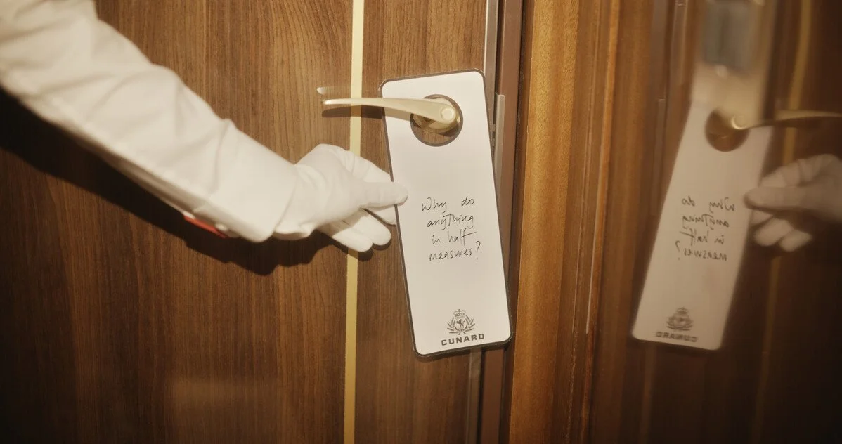 A person in a white glove placing a white door hanger with handwritten text on a hotel door handle. The door hanger reads, "Why do ants run in front of me?" and has the Cunard logo at the bottom. A mirror shows the reflection of the hanger and the door handle.