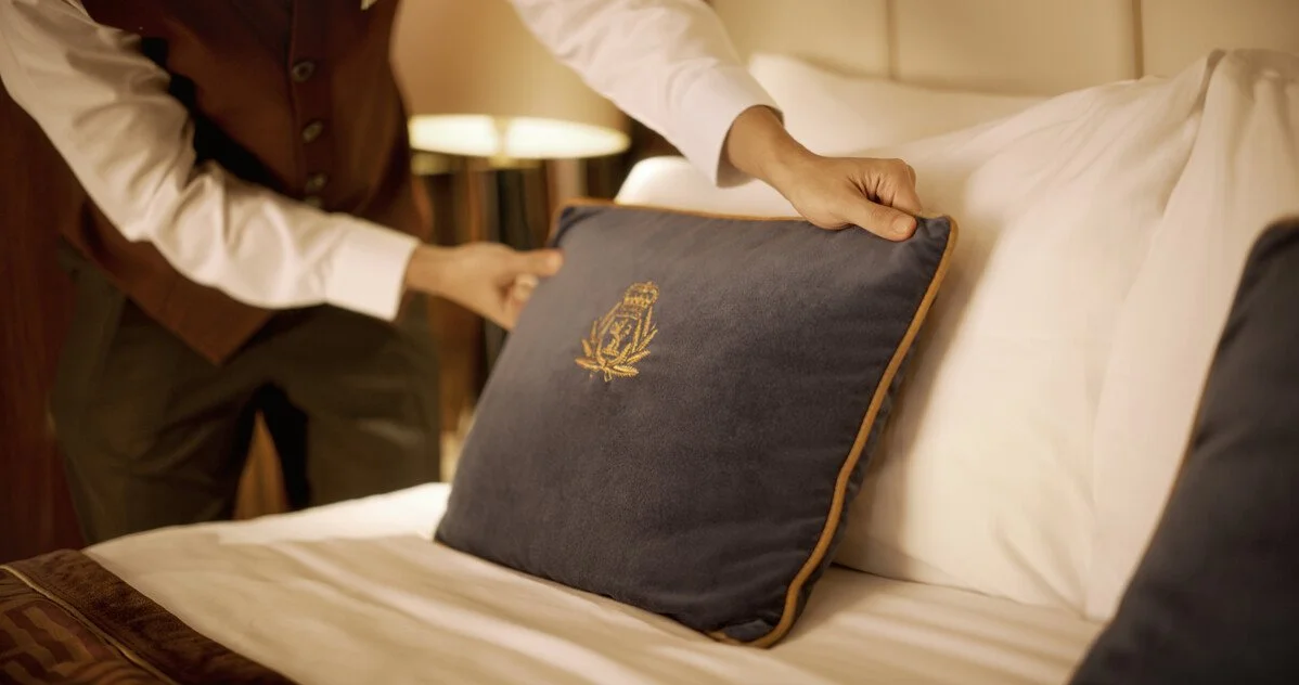 Cleaner arranging a blue decorative pillow embroidered with a crest on a hotel bed.