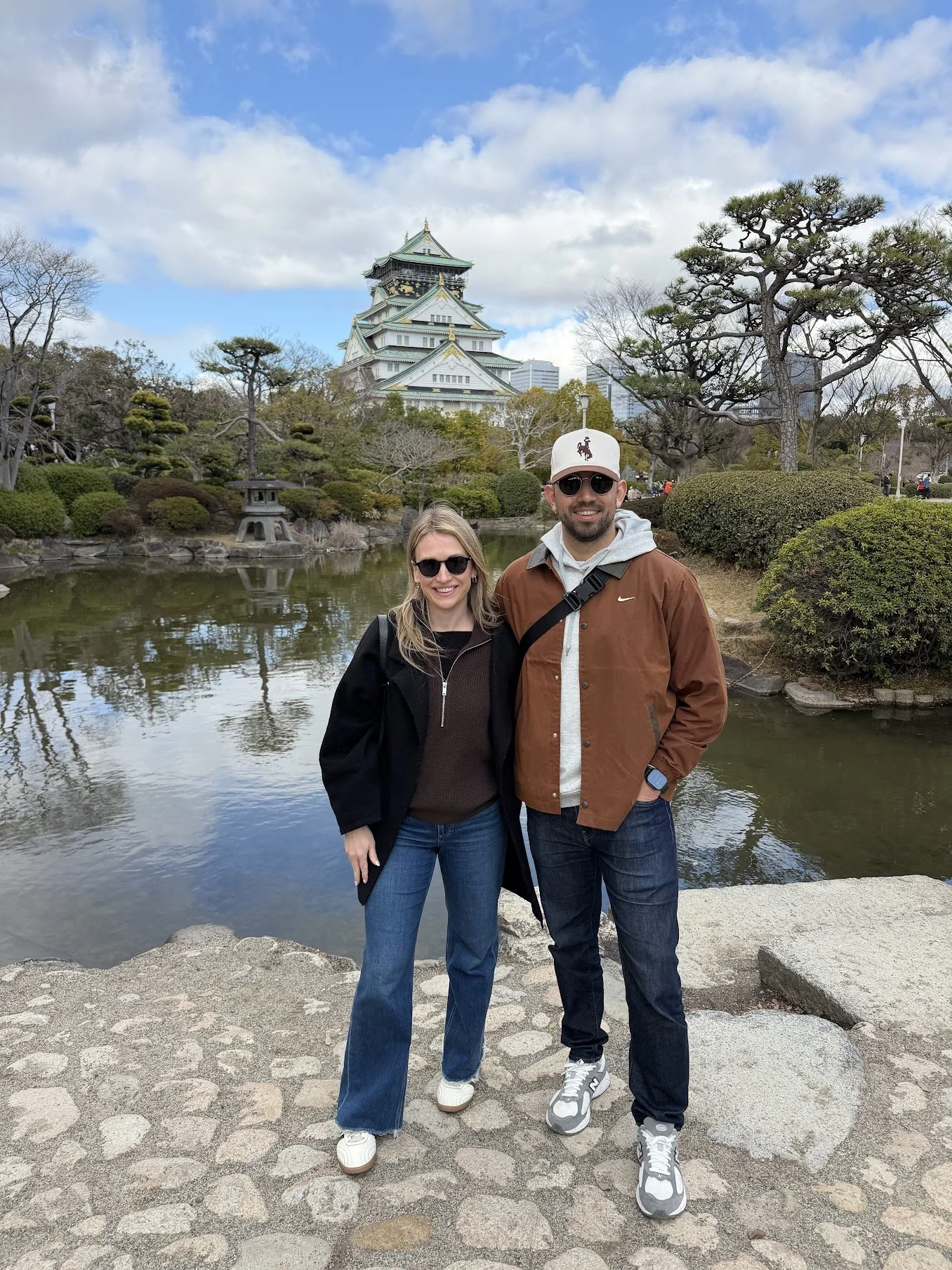 A smiling couple posing outdoors near a pond with a traditional Japanese castle in the background. The woman has blonde hair, wearing sunglasses, a black jacket, and jeans. The man has a beard, wearing sunglasses, a white cap, a brown jacket, and dark jeans.