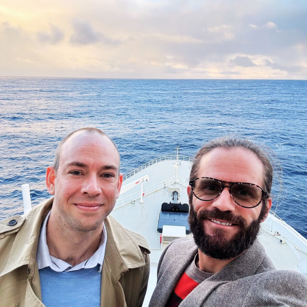 Two men taking a selfie on the deck of a boat out at sea during sunset.