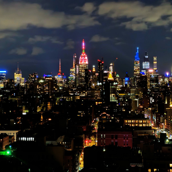 Nighttime view of New York City skyline with illuminated buildings, including the Empire State Building lit in red and blue, and other skyscrapers under cloudy sky.