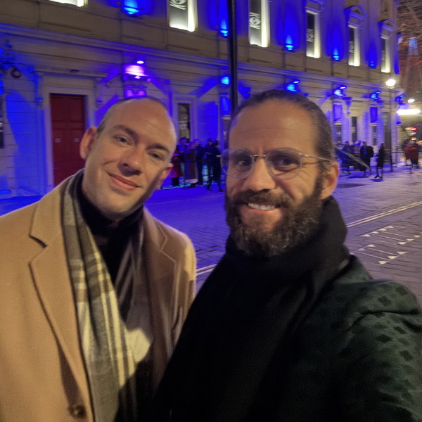 Two men taking a selfie on a city street at night, with a building lit up in blue lights in the background.