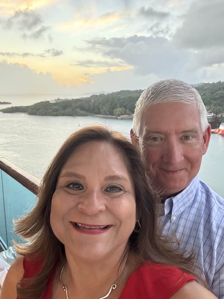 A smiling woman in a red dress and a man in a blue checkered shirt taking a selfie on a balcony overlooking a body of water and a lush, green landscape during sunset.