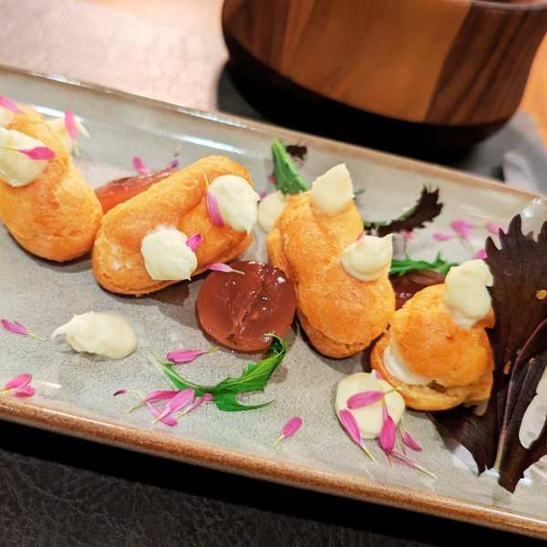 Fried appetizer with puffy, golden-brown shells filled with creamy filling, topped with dollops of white sauce, garnished with edible purple flower petals and greens, on a rectangular plate.