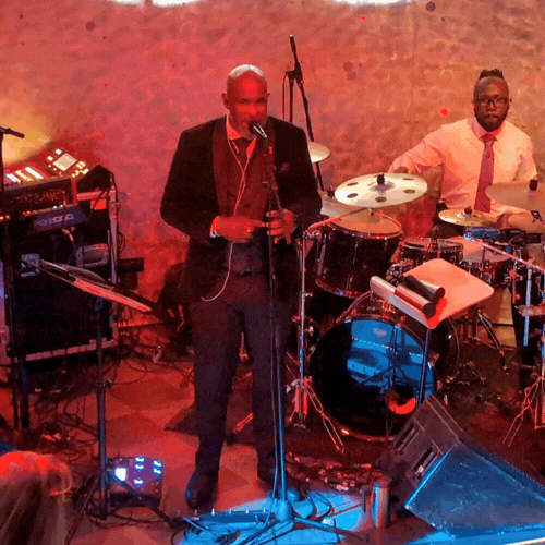 Musicians performing live on stage, with one man singing into a microphone and another playing drums, in a dimly lit venue with brick walls.