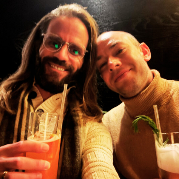 Two men smiling and taking a selfie at a bar or restaurant, each holding a drink, with dark background and warm lighting.