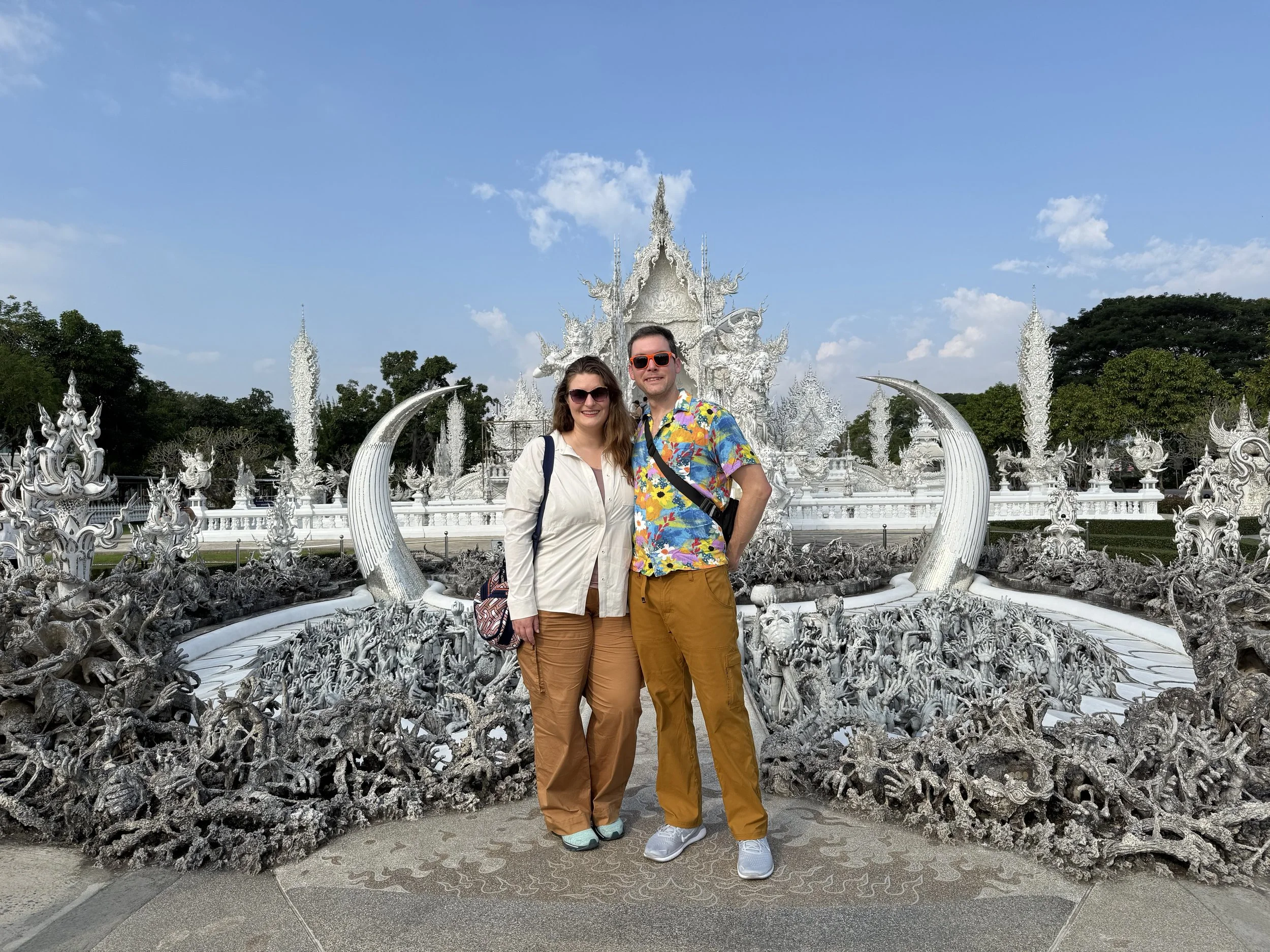 A smiling couple stands in front of a white ornate temple with intricate carvings, surrounded by lush green trees, under a blue sky with some clouds.