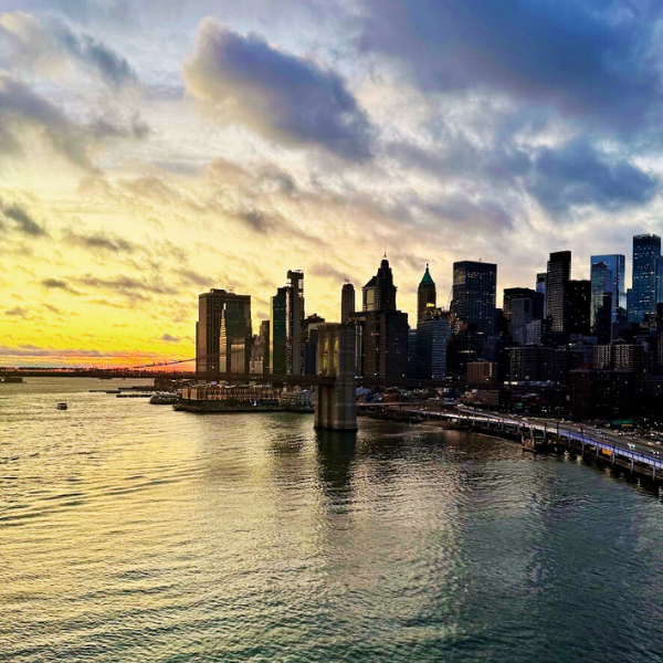 View of Manhattan skyline at sunset with water in the foreground and partly cloudy sky.