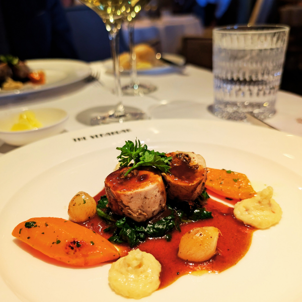 A plated gourmet meal consisting of two pieces of seared meat topped with a sprig of parsley, served with roasted carrots, dollops of mashed potatoes or a similar creamy side, and a green vegetable, all drizzled in a rich sauce on a white plate in a 