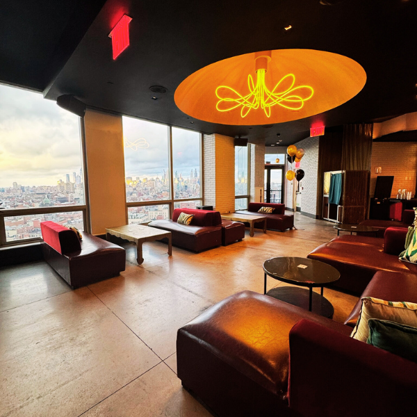 Upscale lounge area with large windows overlooking a city skyline, featuring modern red sofas, a unique yellow neon light fixture on the ceiling, and balloons in the background.