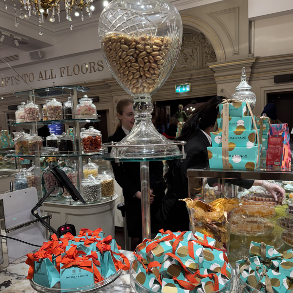 Large glass candy jar filled with mixed nuts on a tall stand at a candy shop. Background shows jars of sweets and shoppers.