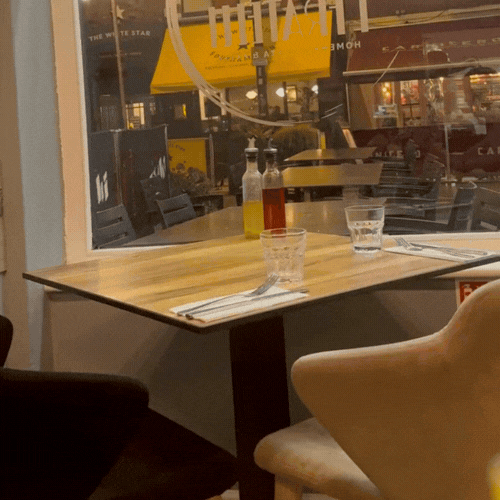 Empty restaurant table set with two glasses, a bottle of oil, a bottle of vinegar, and a laminated menu, next to a large window with a view of an outdoor dining area.