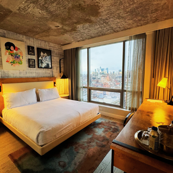 Hotel room with a large bed, artwork on the wall, a window with city view, and a wooden desk with a lamp and personal items.