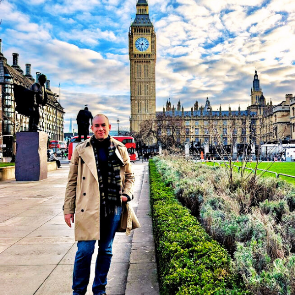 A man standing on a sidewalk in front of Big Ben and the Houses of Parliament in London, with a garden and statues nearby, under a partly cloudy sky.