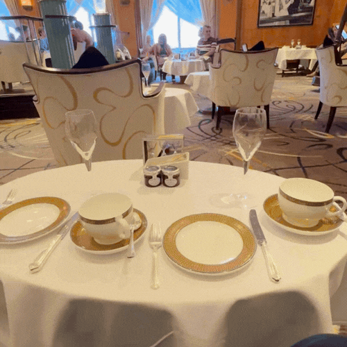 A round table set for two in a restaurant with white and gold china, glasses, and silverware, with a decorated paper napkin holder in the center and a larger glass of water or wine. In the background, other tables and chairs are visible, with patrons
