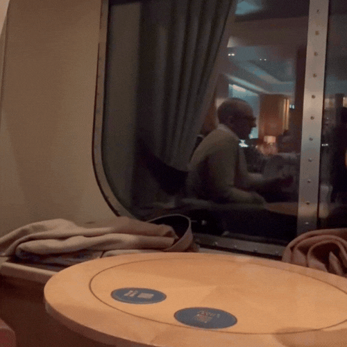 A person sitting at a table in an airport lounge, seen through a window, with a curtain and reflection visible in the window.