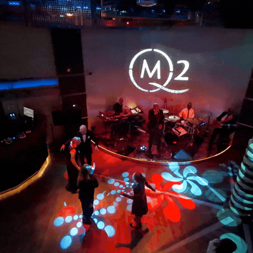A live band performing on stage at an indoor event with a logo that reads 'M 2' projected on the wall behind them, with people dancing or talking in front of the stage and colorful lighting on the floor.