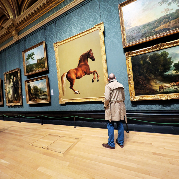 A person with a bald head and beige trench coat looking at a large painting of a jumping horse in an art gallery with ornate gold frames on the wall.
