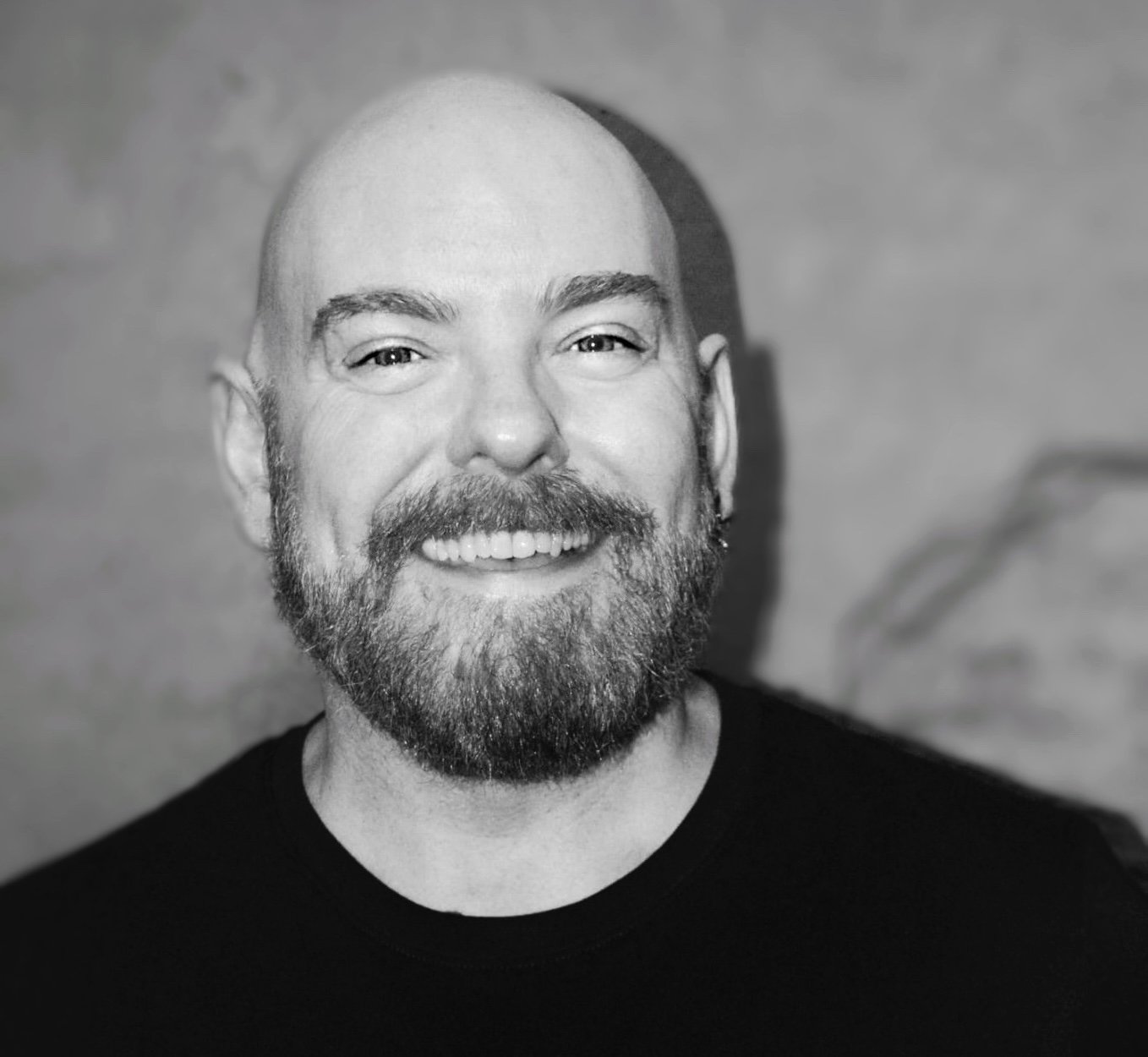 A black and white photo of a smiling man with a beard, mustache, and a shaved head, wearing a black shirt. The background is plain and slightly blurred.