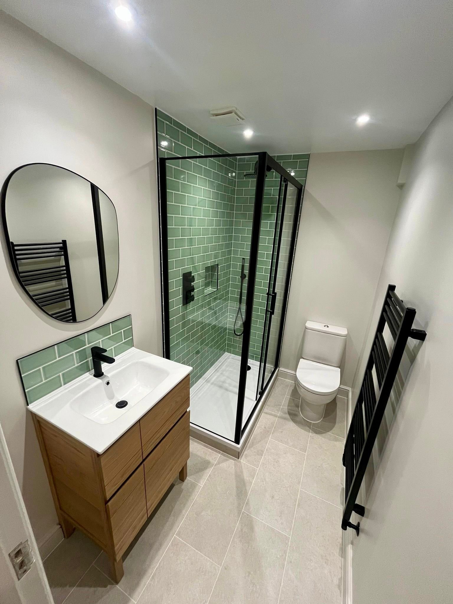 Modern bathroom with a green tiled shower, white toilet, wooden vanity with black faucet, round mirror, black towel rack, and beige tiled floor.