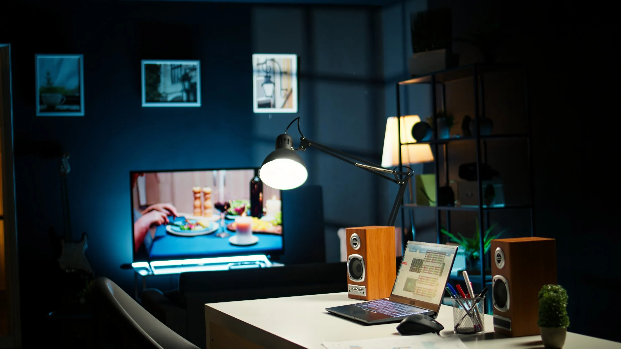 Home office with a desk, laptop, computer speakers, potted plant, and a monitor showing a dinner scene. A wall-mounted light, framed pictures, and a shelving unit are in the background.