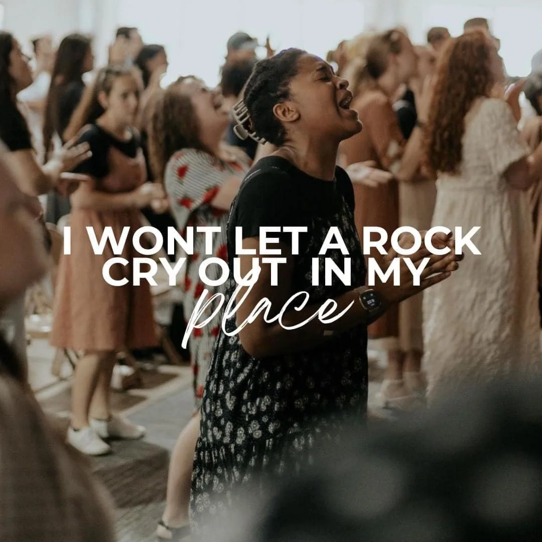 A group of people at a religious or worship gathering, with a prominently displayed text saying, "I WON'T LET A ROCK CRY OUT IN MY PLACE."
