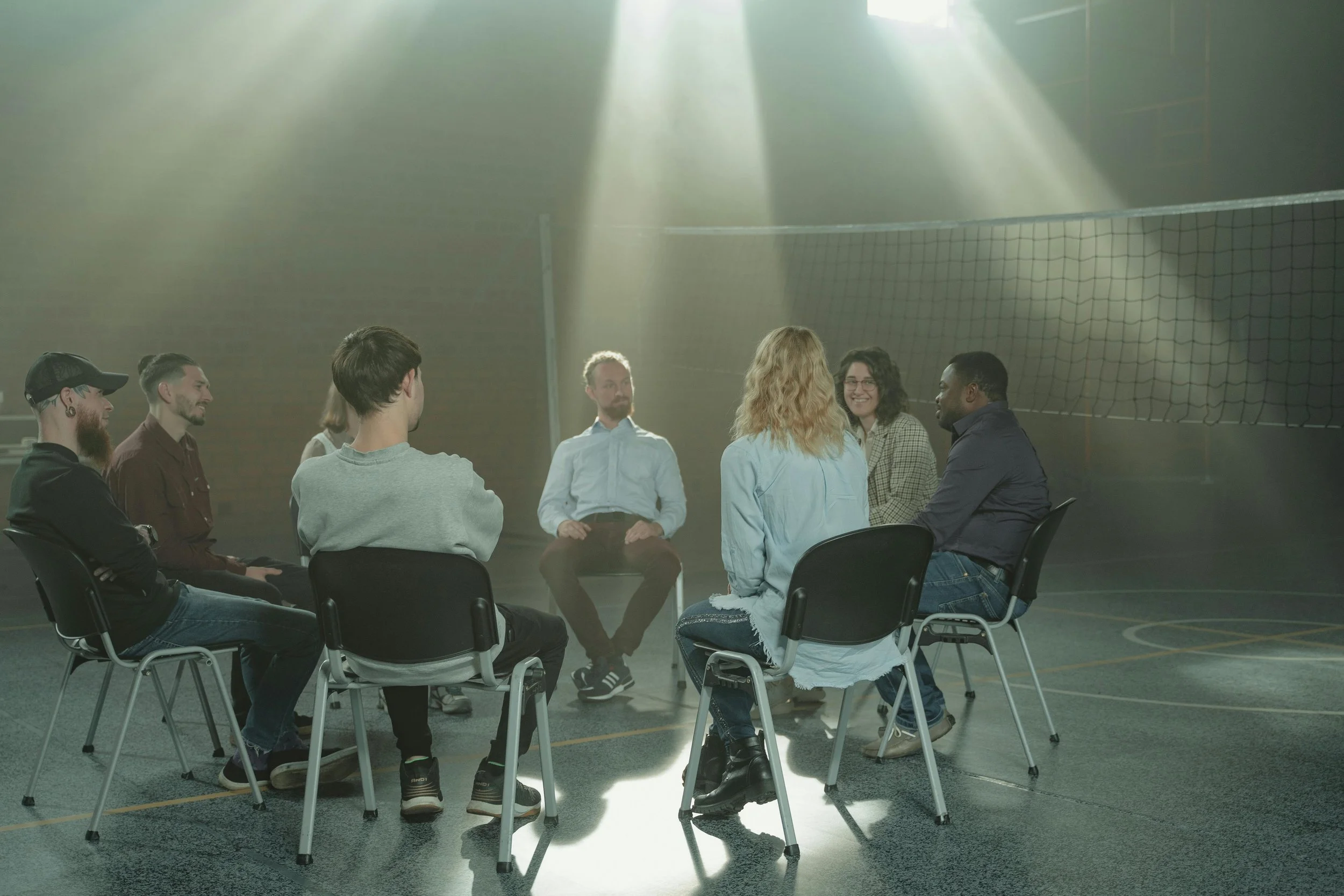 A group of people seated in a circle having a discussion in a dimly lit room with a net in the background. Sunlight streams through the windows, creating beams of light on the floor.