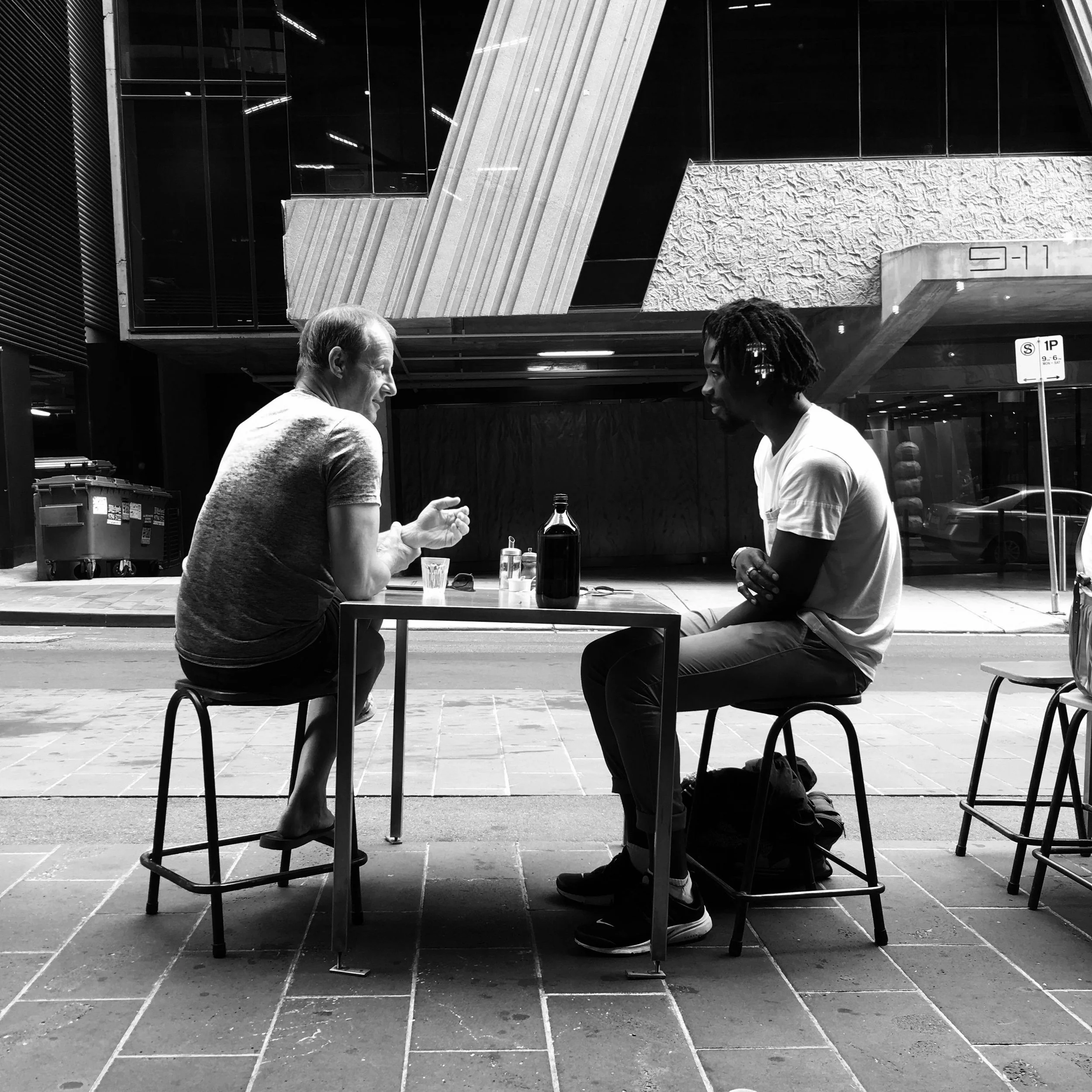 Two men sitting at an outdoor table having a conversation in an urban setting, with a large building in the background. They are seated on stools with drinks on the table, and the image is in black and white.