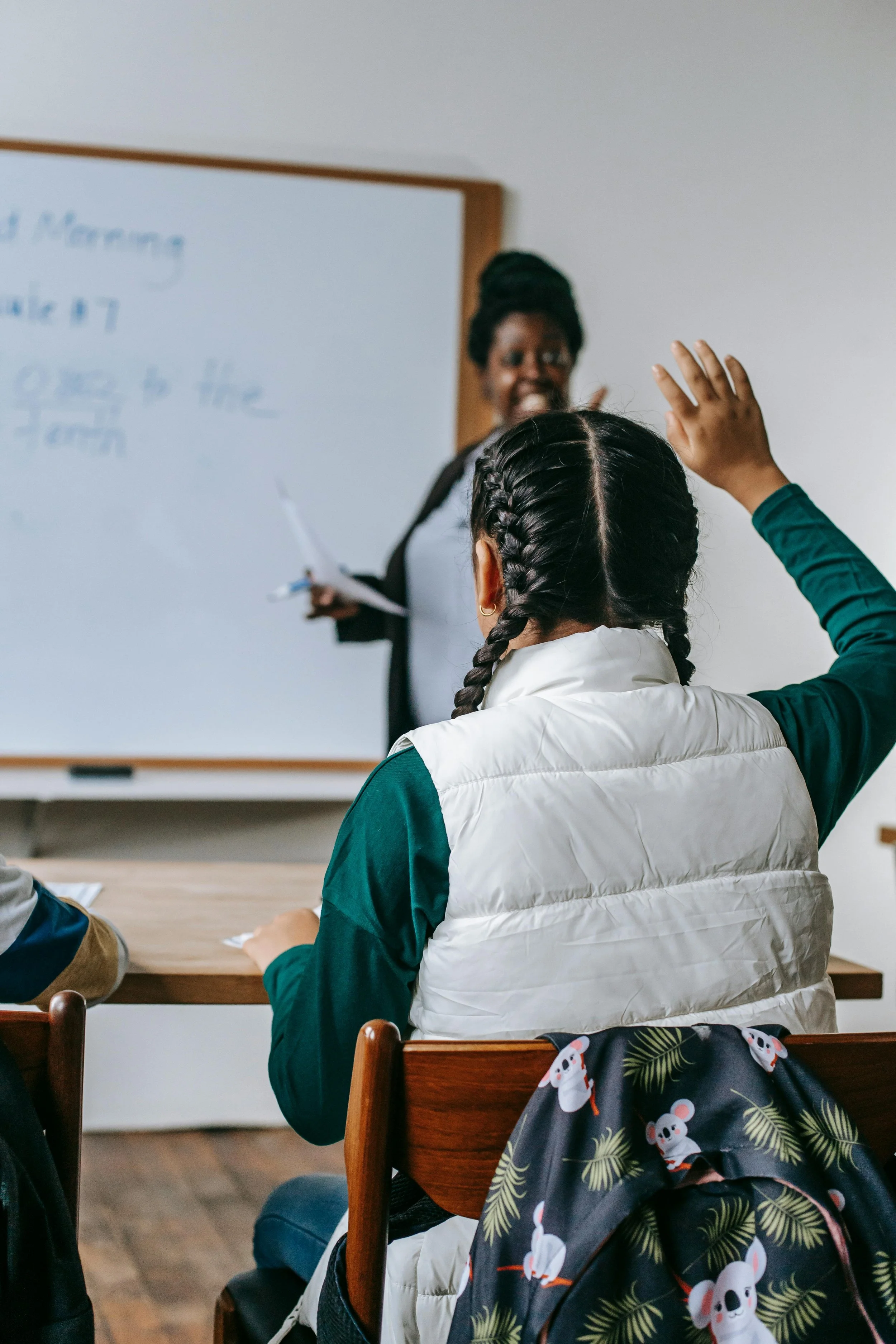 A student raising their hand in a classroom while a teacher holds papers in front of a whiteboard.