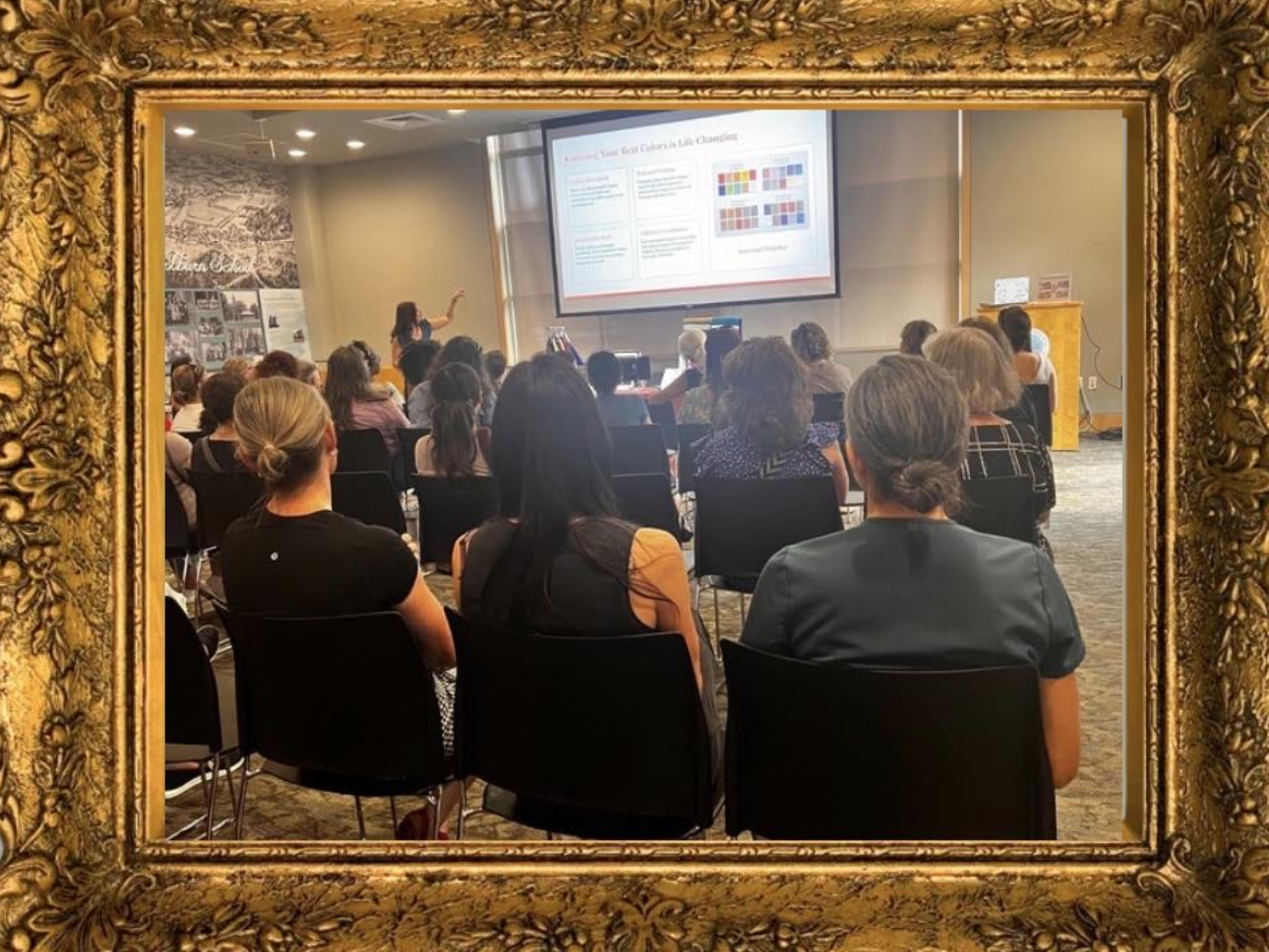 Group of women attending a presentation or workshop in a conference room, seen through an ornate gold picture frame. The presenter is pointing at a large screen with a colorful slide.