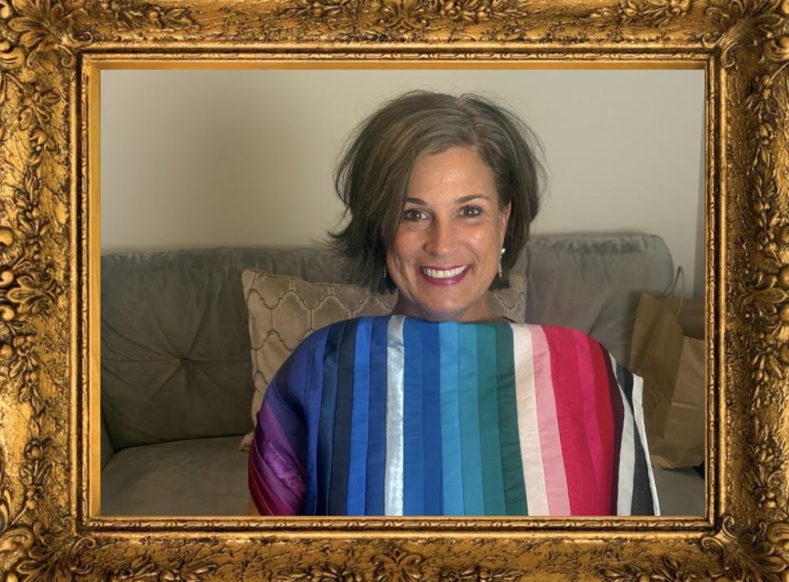 A woman with short brown hair, wearing a colorful rainbow-striped shawl, is smiling and sitting on a beige couch in a room with plain walls. The image is framed with an ornate, gold decorative frame, giving the appearance that her photo is in a picture frame.