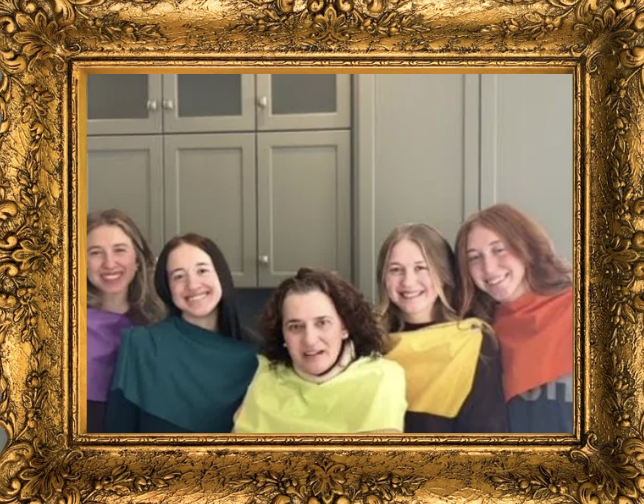 Group of five women smiling, wearing colorful shawls, standing in front of a gray cabinet, framed by an ornate gold frame.