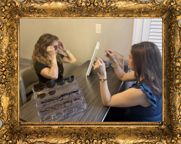 A woman fitting glasses on a girl at a table with a display of eyeglasses, framed by an ornate gold frame.