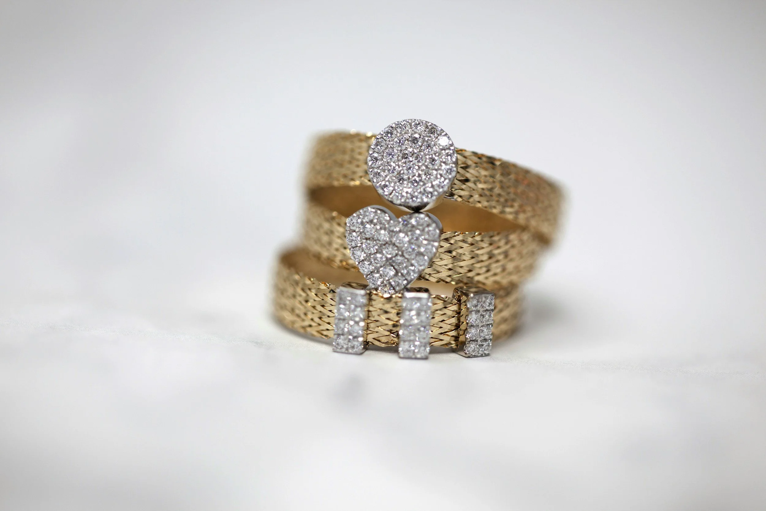Close-up of a gold ring with a round face covered in small diamonds, placed on a textured gold band, with matching diamond earrings in front, all reflected on a glossy surface.