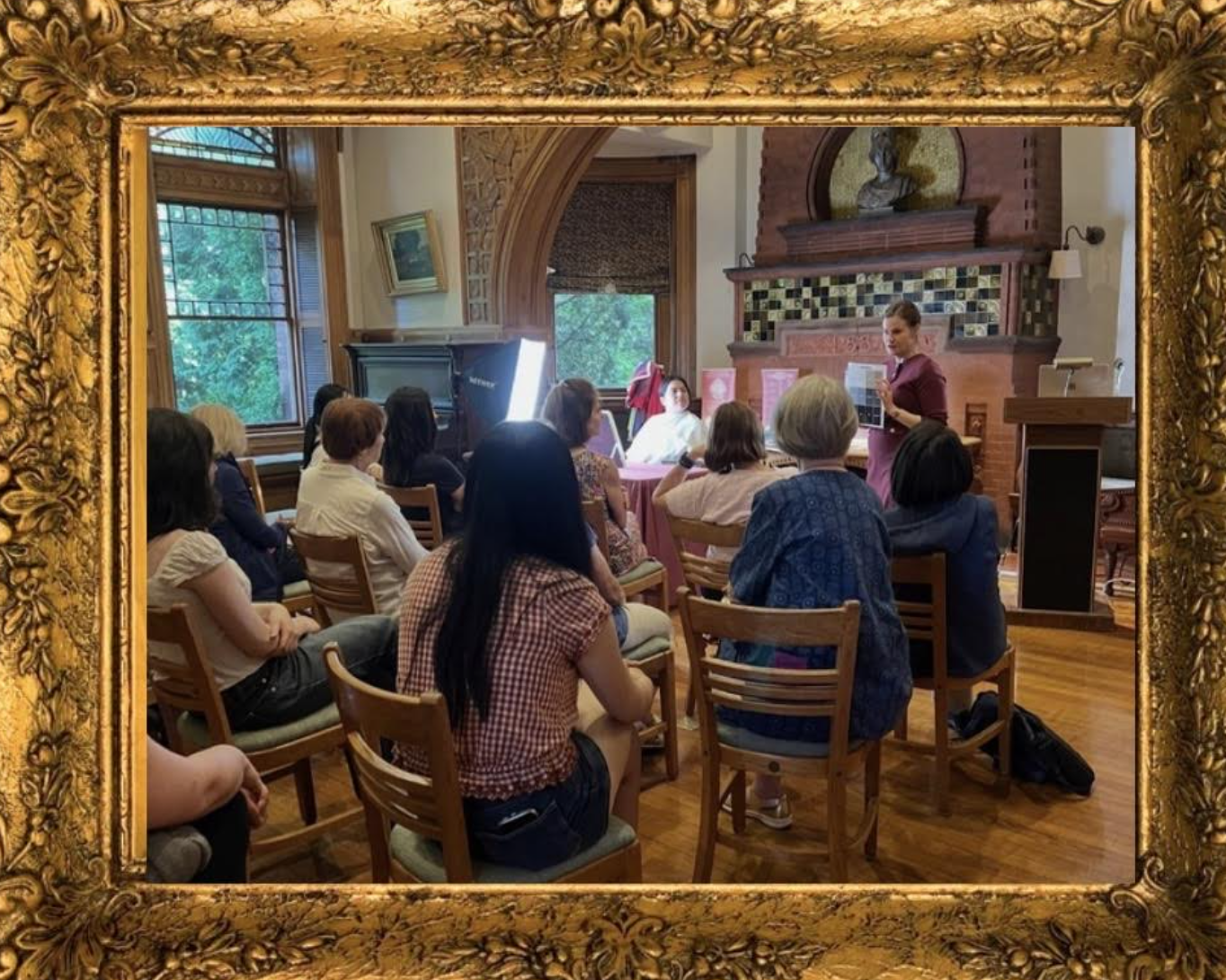 A group of people seated in a room with wooden floors and large windows, attending a presentation or lecture by a woman standing near a podium. The room has a brick fireplace and artwork on the walls.
