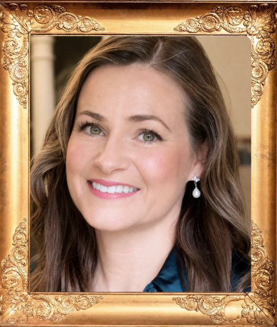 A woman with long brown hair and green eyes, smiling, wearing pearl earrings and a dark top, framed by an ornate gold border.