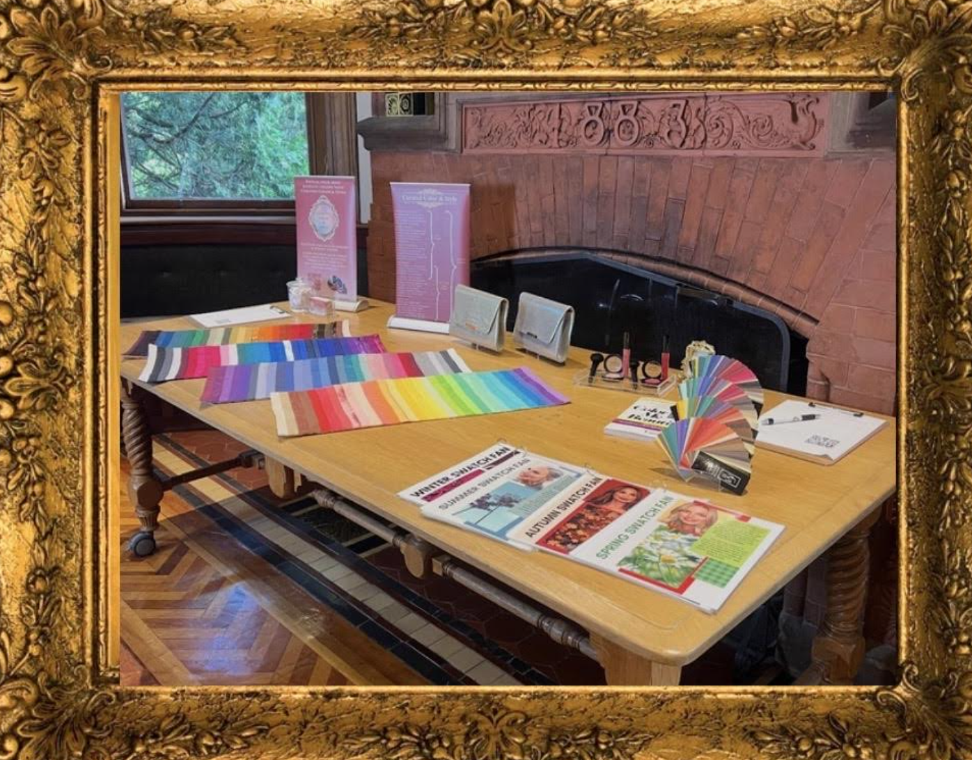 A wooden table with colorful fabric swatches, pamphlets, brochures, and paint samples. Behind the table is a brick wall fireplace and a window showing green trees outside. The table is in a richly decorated room with a gold ornate frame border.